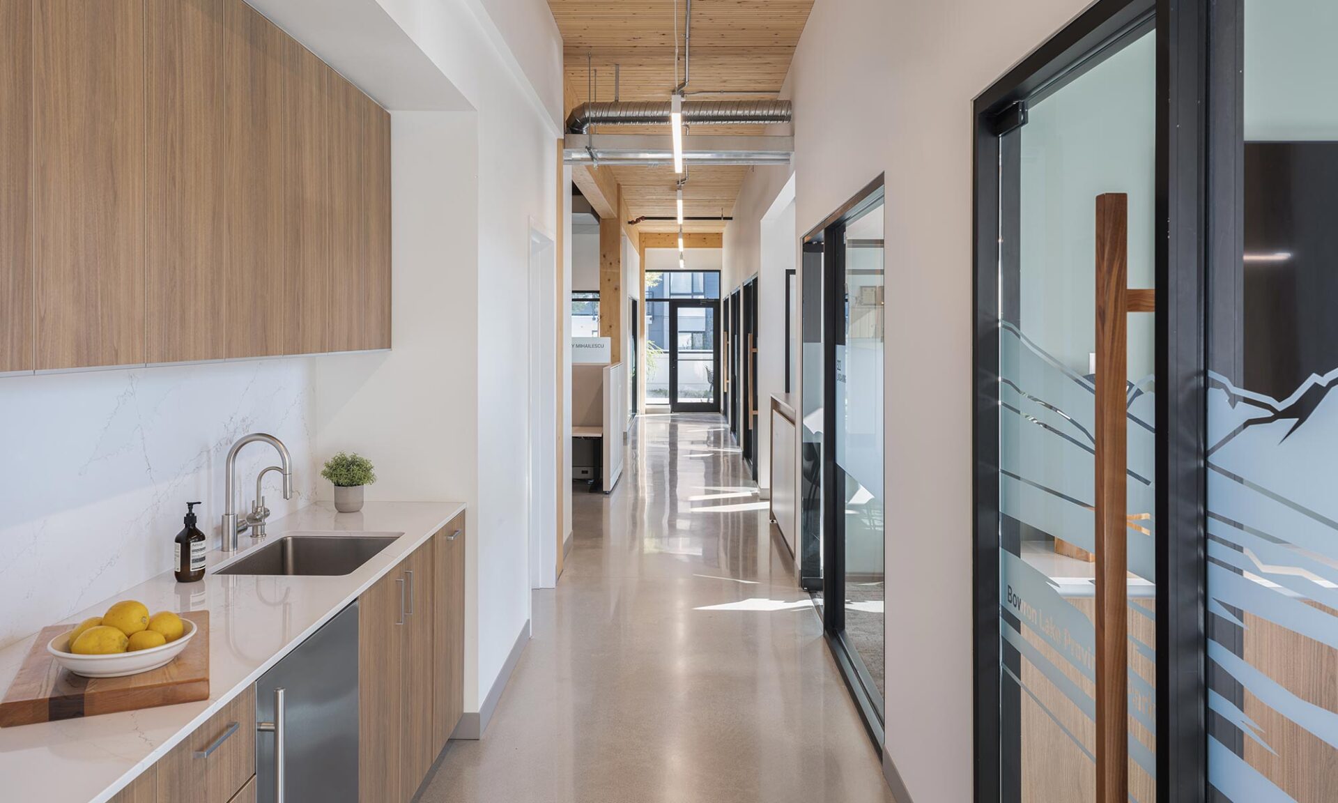 Bright office corridor blending natural daylight with suspended architectural lighting below exposed wood ceiling.