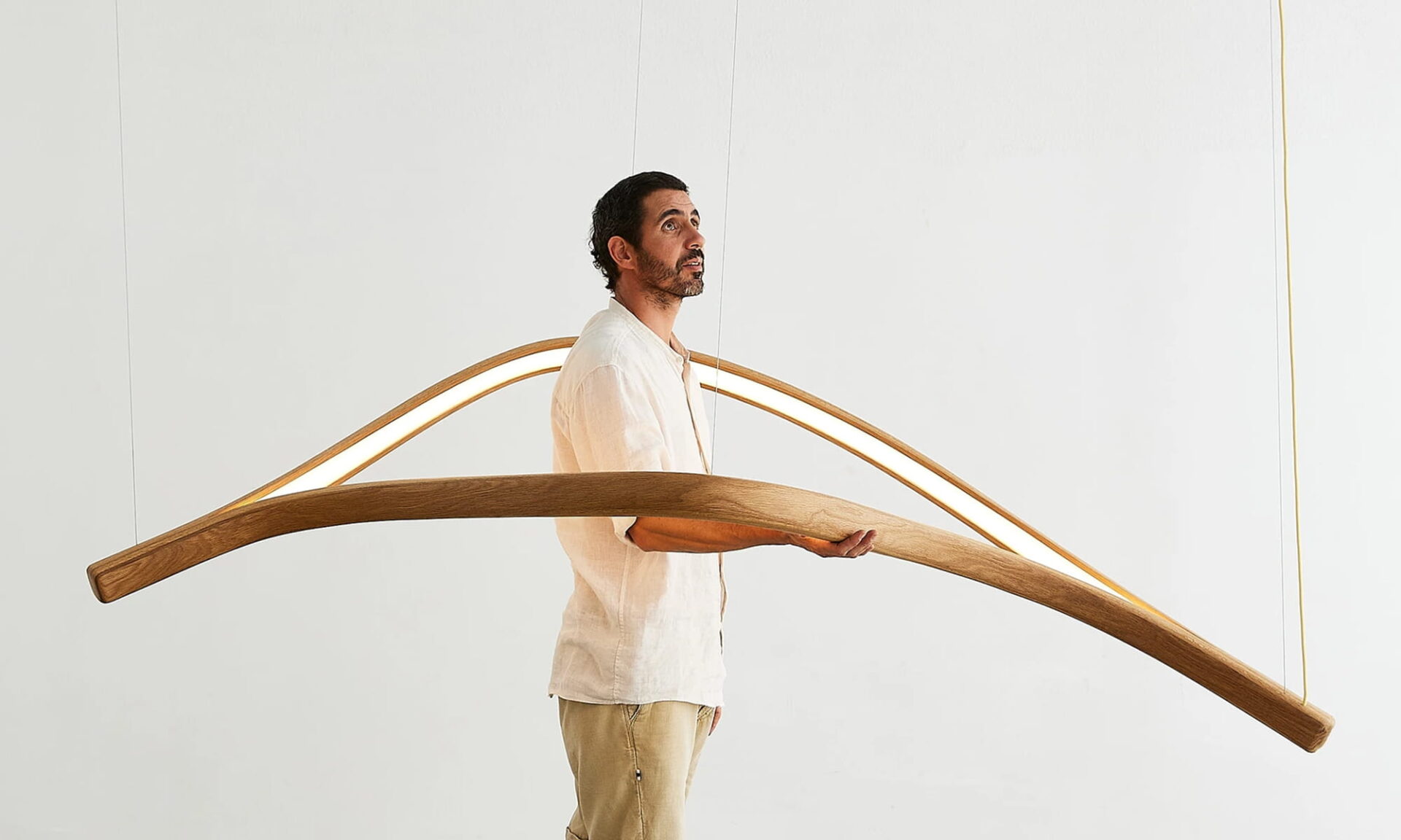 Poémia founder Ricardo Costa holds a sculptural wooden luminaire, showing the handcrafted, flowing wave-like form inspired by the ocean.