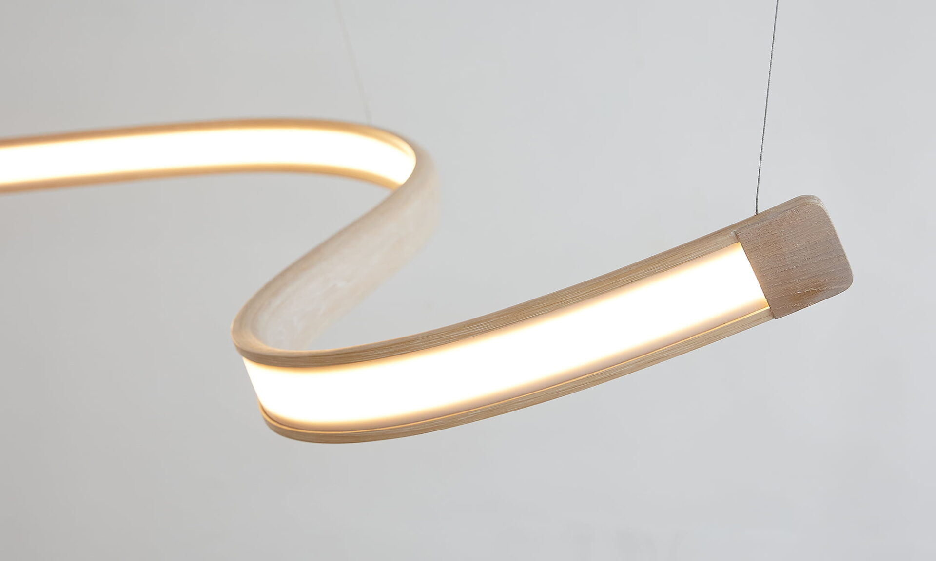 Poémia Brisa ceiling lamp in ash wood, a sculptural pendant with flowing, wave-like curves, shown illuminated in a modern interior.