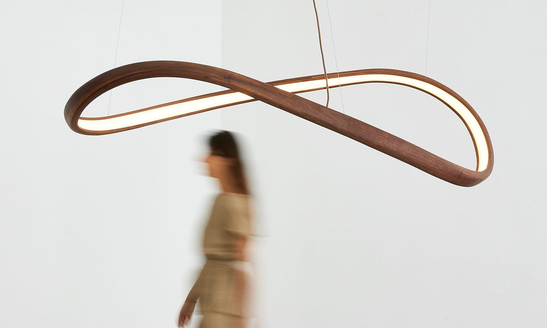 Poémia Horizonte ceiling lamp in walnut wood, a sculptural pendant with an infinity shape, shown softly illuminated in a modern interior with a person walking below it.