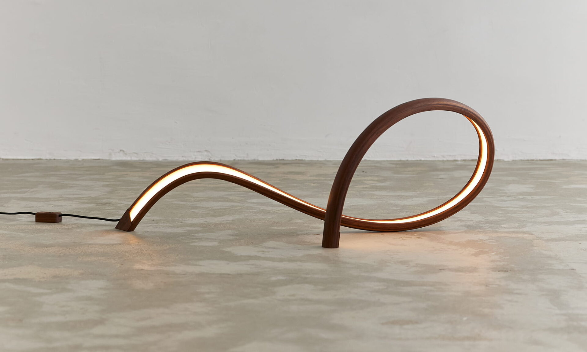 Poémia Maré table lamp lit on the floor, a sculptural walnut wood luminaire with a looping curve inspired by the ocean waves.