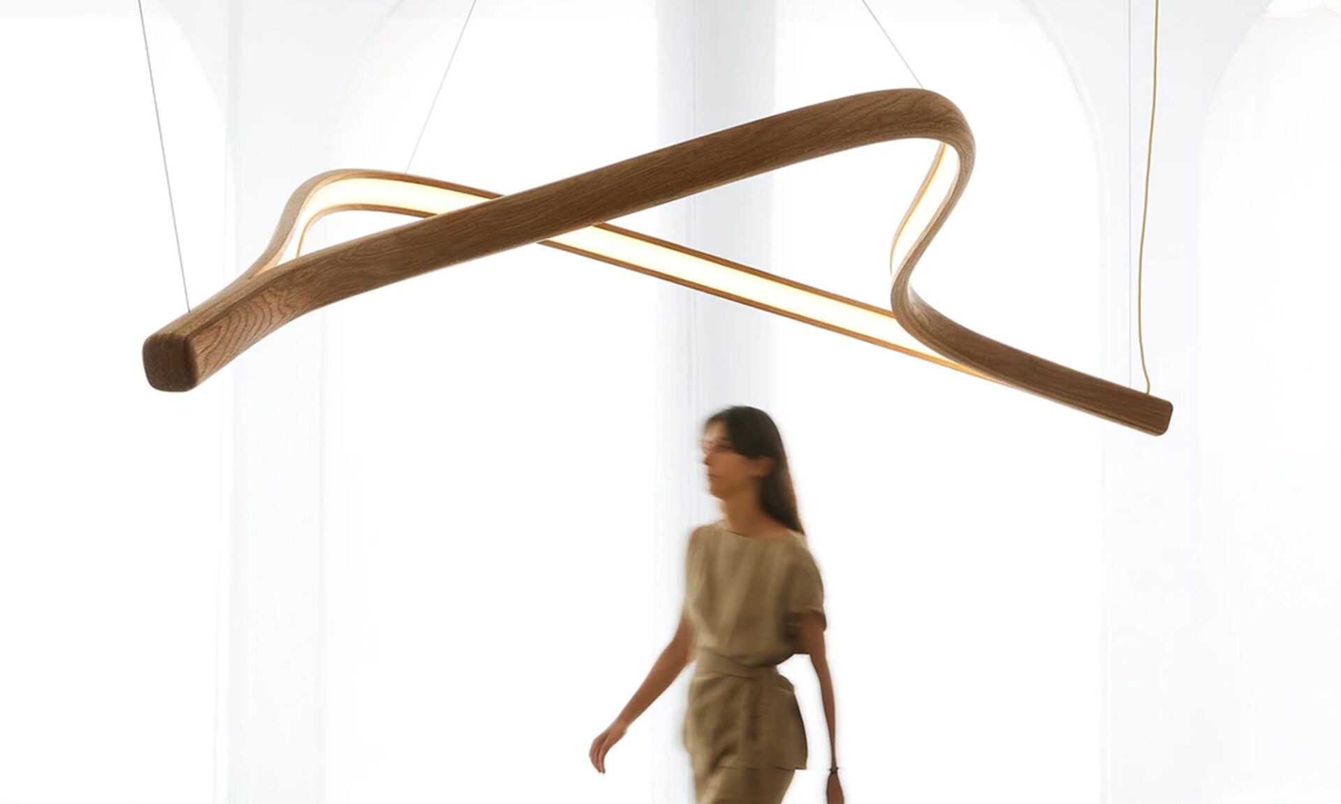 Poémia Pérola ceiling lamp in oak wood, a sculptural pendant with smooth, wave-like curves inspired by the sea, shown softly illuminated in a modern interior with a women walking underneath it.