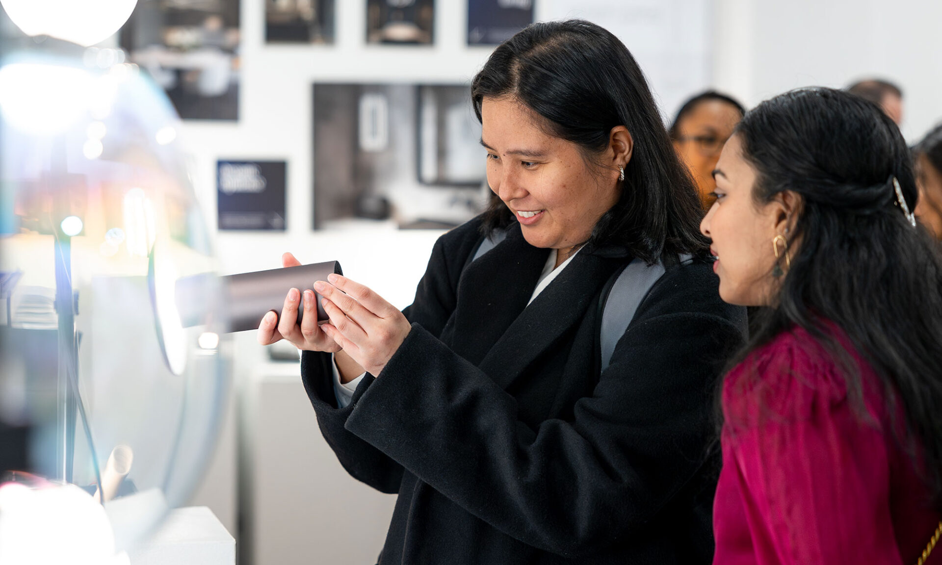 Two guests at Lumorphéa in Vancouver examine an Artemide Takku portable lamp up close beside a glowing display, with additional exhibit lighting and gallery displays in the background.
