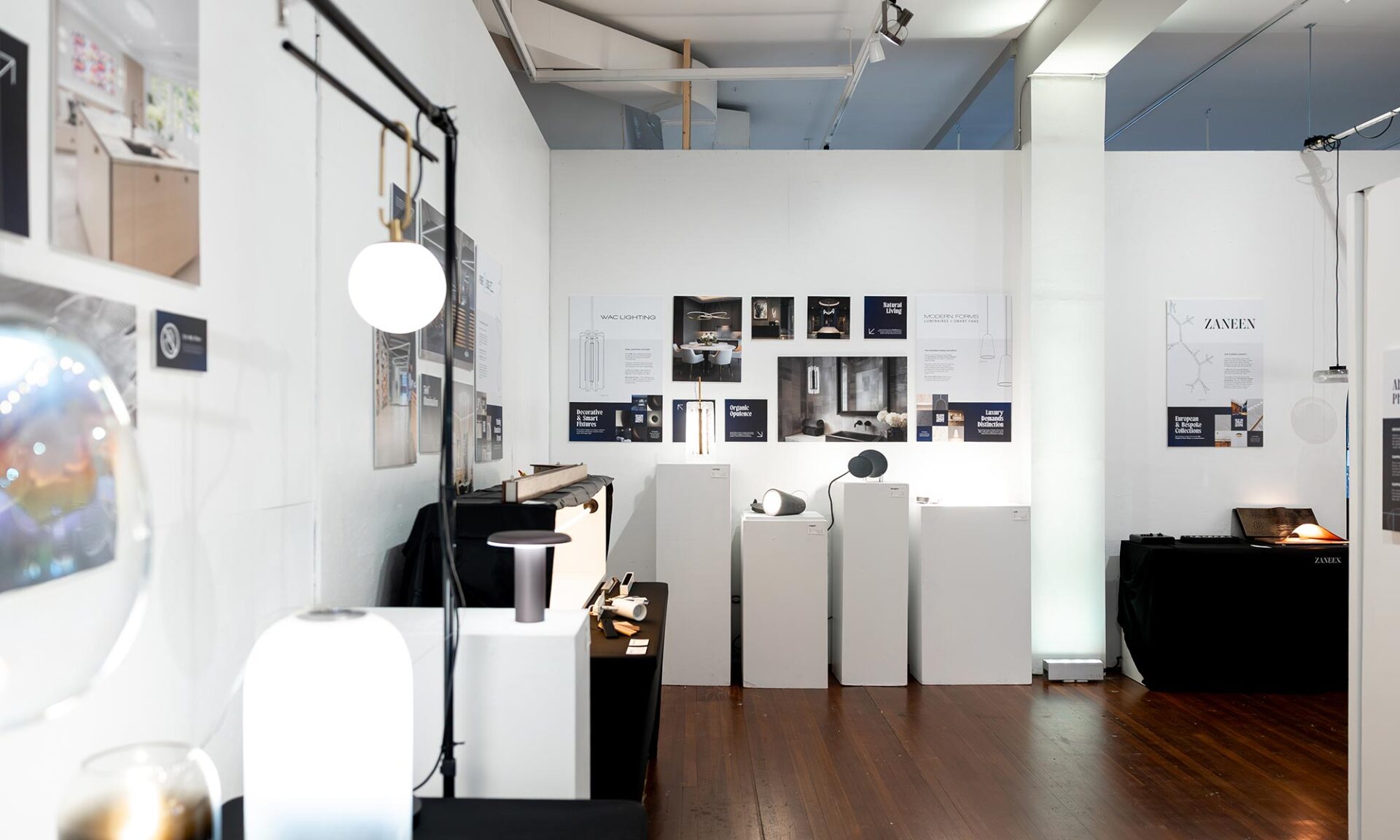 Wide interior photo of the Lumorphéa exhibition at The Gallery George in Vancouver, showing minimalist lighting fixtures on display tables and plinths, with brand panels and gallery walls in the background.