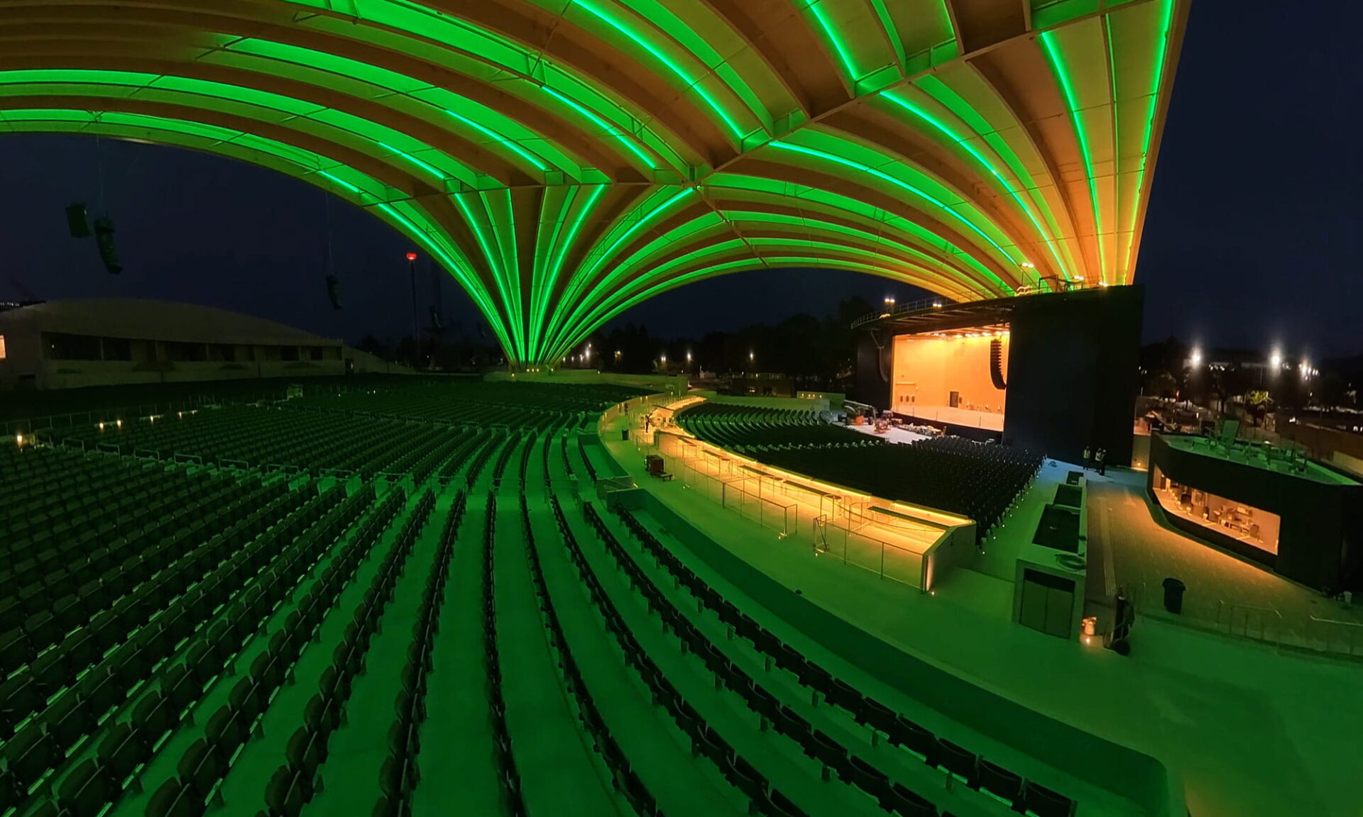 Angled interior nighttime view inside the PNE Amphitheatre Freedom Mobile Arch showing the mass timber roof illuminated in green, with parts of the stage and seating visible.