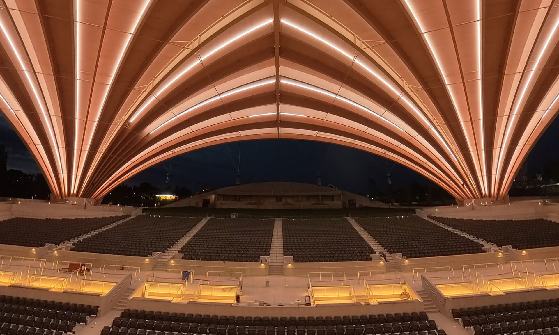 Interior view toward seating under the PNE Amphitheatre Freedom Mobile Arch at night, showing neonVIEW lines along the curved timber beams.
